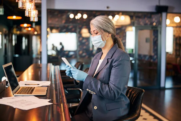 Charmant zakenvrouw in medisch masker met behulp van smartphone in cafe — Stockfoto