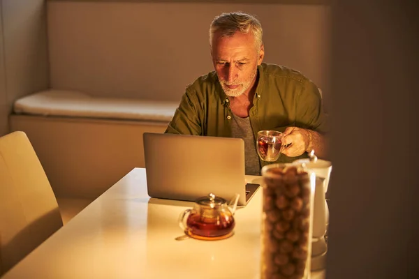 老先生在笔记本电脑上工作时喝茶 — 图库照片