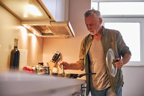 Старший джентльмен снимает крышку с сковороды на плиту — стоковое фото