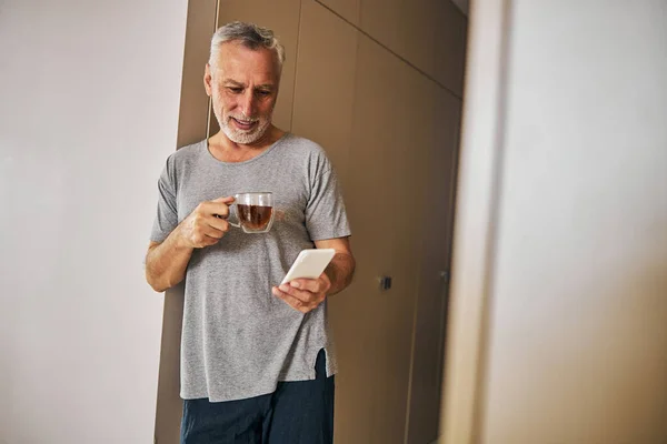 Senior άνθρωπος έλεγχο smartphone του, ενώ πίνοντας τσάι στο σπίτι — Φωτογραφία Αρχείου