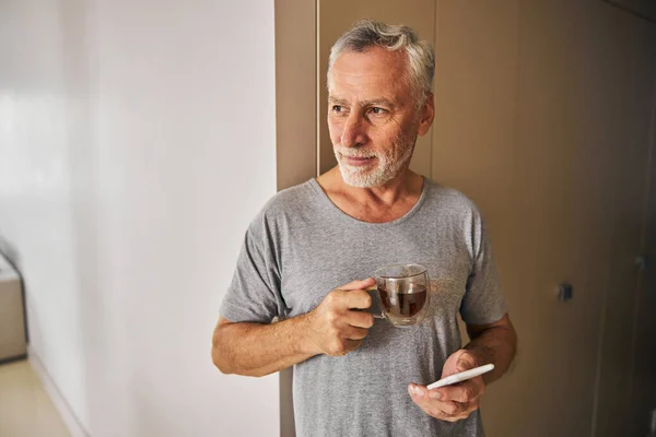 Aged gentleman having tea at home and holding his phone — Stock Photo, Image