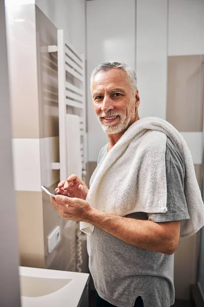 Χαρούμενος ηλικιωμένος κύριος με ένα smartphone κοντά στο νιπτήρα του μπάνιου — Φωτογραφία Αρχείου