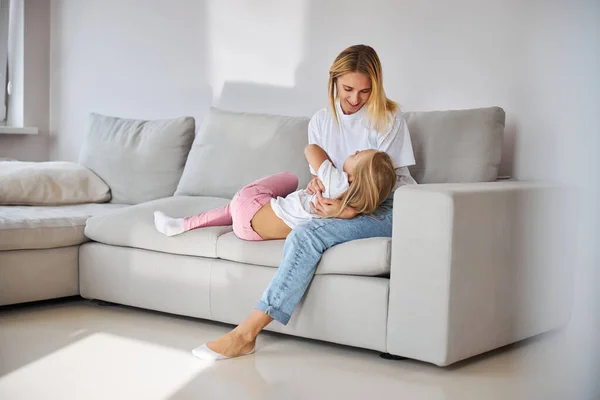 Elegante madre divirtiéndose con su hija en el sofá en la habitación gris — Foto de Stock
