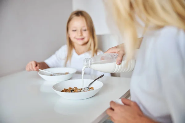 Donna adulta versando il latte per la colazione mentre è seduta con la bambina in cucina a casa — Foto Stock