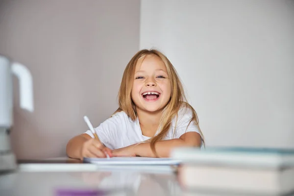 Happy laughing little girl sitting at the white desk and doing writing exercises in book — Stock Photo, Image