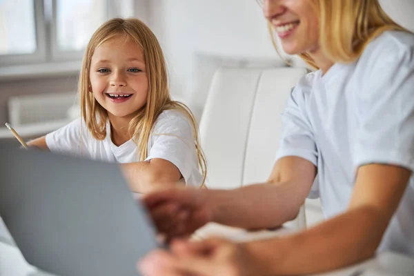 Feliz hija sonriente mirando a la computadora portátil de pantalla mientras está sentada cerca de la madre —  Fotos de Stock