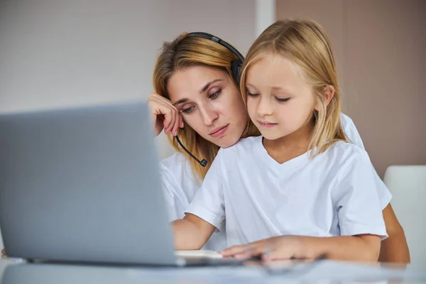 Sad mother in headphone spending time with her daughter while sitting at the desk in room inside — Stock Photo, Image