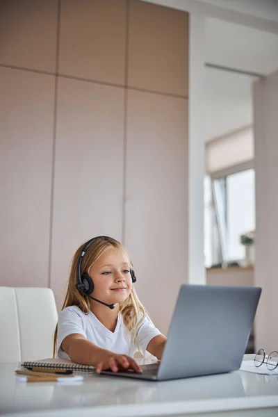 Charming little miss looking to the screen her grey laptop while sitting at the white table in her room — Stock Photo, Image