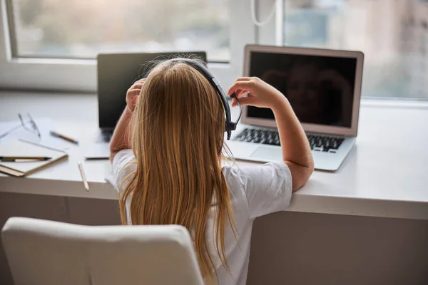 Carino piccola signorina in abiti casual guardando i due schermo i suoi computer portatili mentre seduto al tavolo bianco — Foto Stock