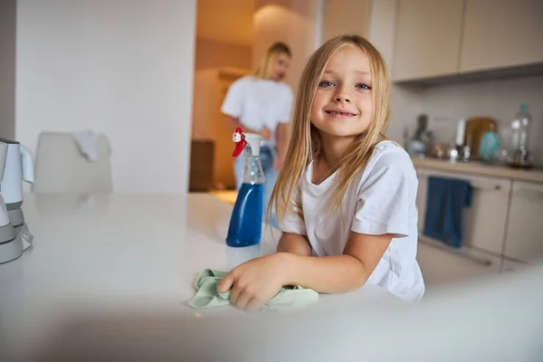 Felice sorridente piccola signorina in piedi al tavolo bianco mentre lo lavava — Foto Stock