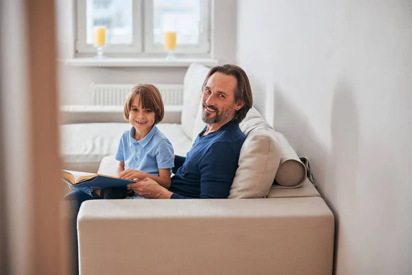 Liebenswertes Kind und sein bärtiger Vater ruhen auf dem Sofa — Stockfoto