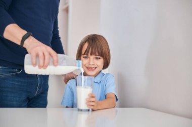Güler yüzlü küçük çocuk babasının süt dökmesi için bardak tutuyor.