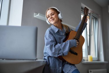 Gitar çalan ve evde dizüstü bilgisayar kullanan hoş bir genç kadın.