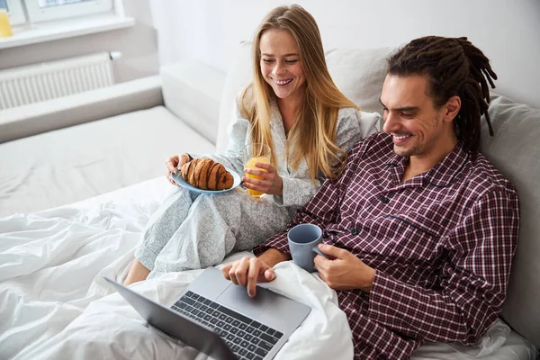 快乐的年轻夫妇使用笔记本电脑，在床上吃早餐 — 图库照片