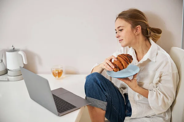 Чарівна молода жінка їсть круасан і використовує ноутбук вдома — стокове фото