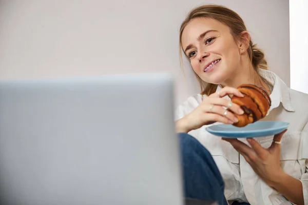 Joyful jonge vrouw met croissant met behulp van notebook thuis — Stockfoto