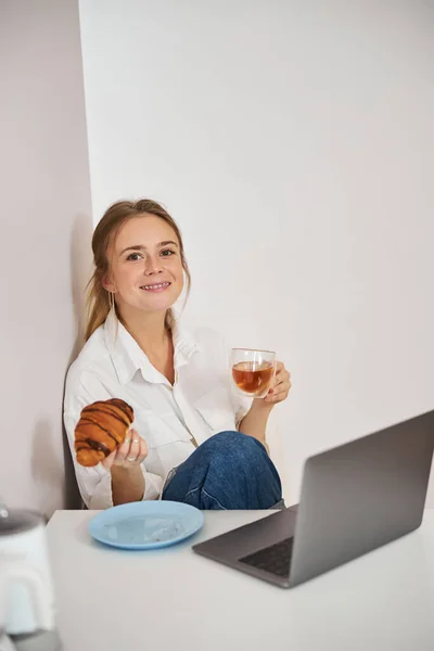 Joven alegre desayunando y usando el portátil en casa — Foto de Stock