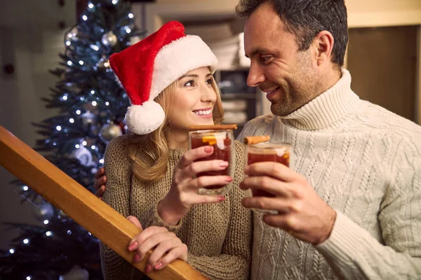 Mulher encantadora em chapéu de Papai Noel olhando para bonito macho em suéter, enquanto eles seguram bebida em braços — Fotografia de Stock