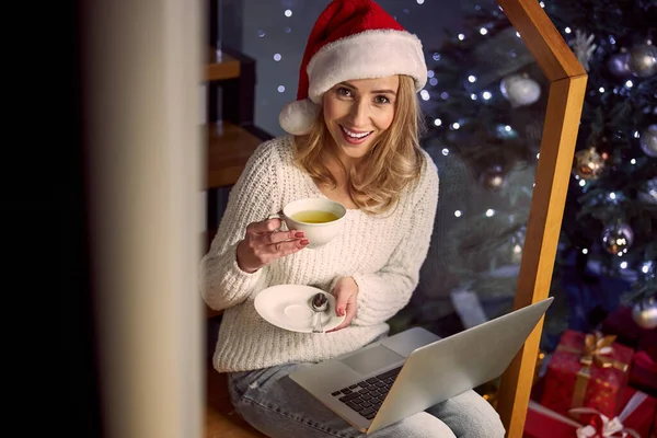 Femme blonde élégante en pull blanc chaud et chapeau de Père Noël tenant boisson dans les mains tout en étant assis sur un escalier en bois dans la maison — Photo