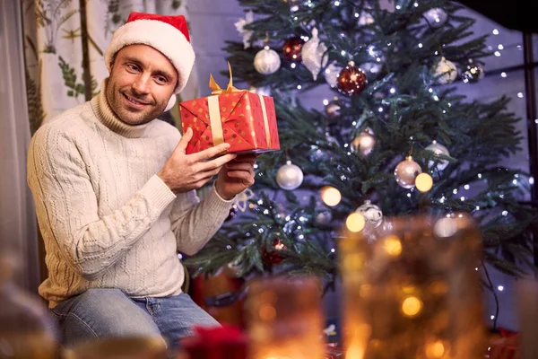 Macho pensativo em suéter e chapéu de Santa segurando presente nos braços no quarto dentro de casa — Fotografia de Stock