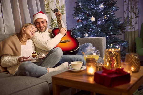 Гарний впевнений чоловік у капелюсі Санта розслабляється з чарівною елегантною дружиною у вітальні на зимові канікули — стокове фото