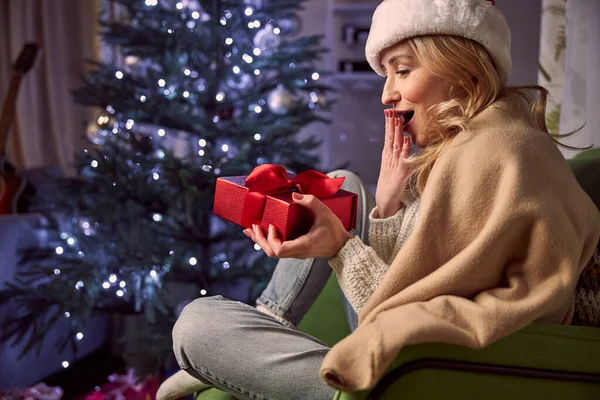 Magnifique femme élégante dans le chapeau Santa regardant vers la boîte cadeau rouge et attendant Noël dans les appartements à la maison — Photo