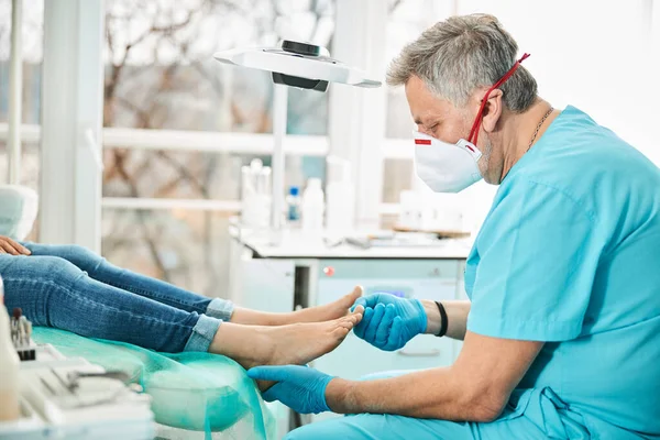 Professionell läkare fotvårdsspecialist i medicinsk mask undersöka fötter och naglar medan patienten sitter i medicinsk stol — Stockfoto