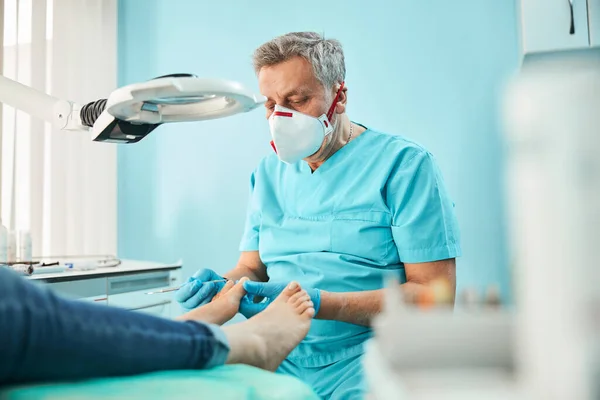 Kiropodist fotvårdsspecialist i blå uniform och handskar med medicinska verktyg i handen under behandlingen förfarande på fötterna — Stockfoto
