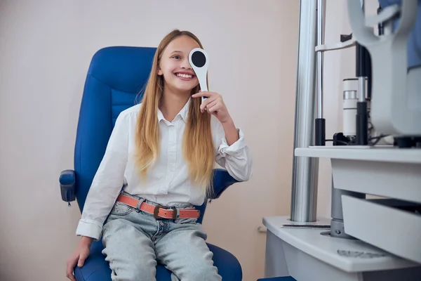 Fröhliches fröhliches weibliches Kind sitzt auf dem Medizinstuhl im Optikerschrank — Stockfoto