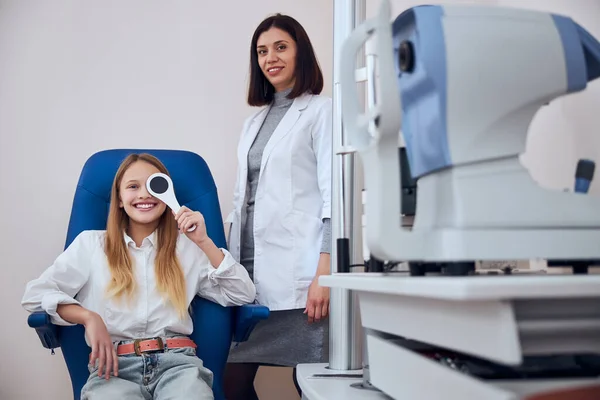 Linda chica adolescente sentada en silla de medicina azul y sosteniendo una herramienta especial en la mano con el médico óptico detrás de ella — Foto de Stock