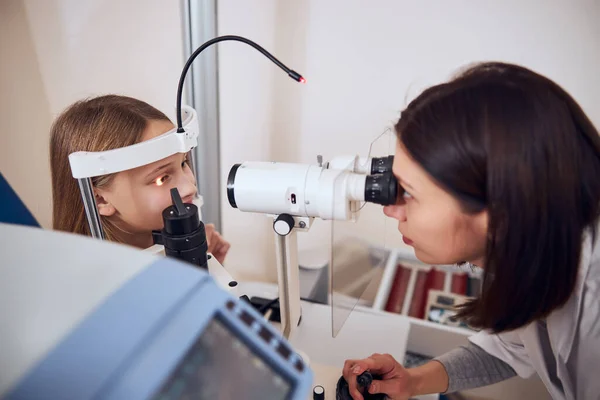 Chica adolescente rubia mirando al médico durante la comprobación de su visión con oftalmoscopio —  Fotos de Stock