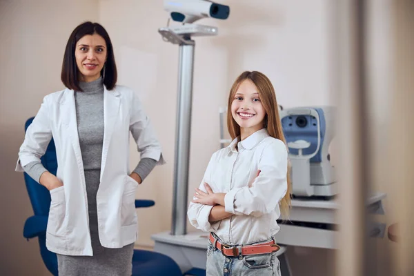 Belle femme enfant et brunette optométriste médecin debout dans le cabinet en face de la machine moderne — Photo