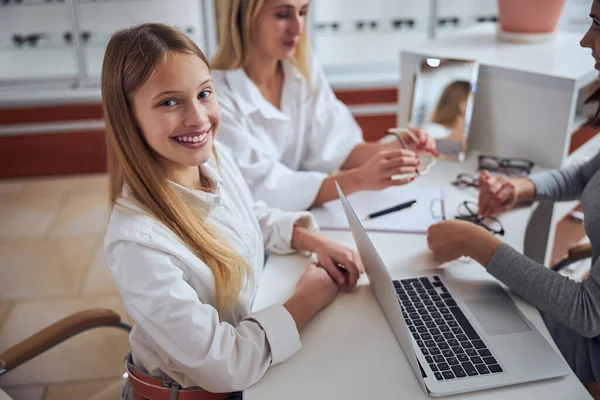 Hermosa niña en camisa blanca mirando a la cámara mientras está sentada en la mesa frente a la computadora portátil —  Fotos de Stock