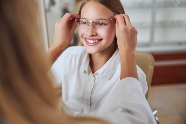 Beautiful friendly young lady looking ahead while unrecognized woman helping to choose glass in room indoors — Stock Photo, Image