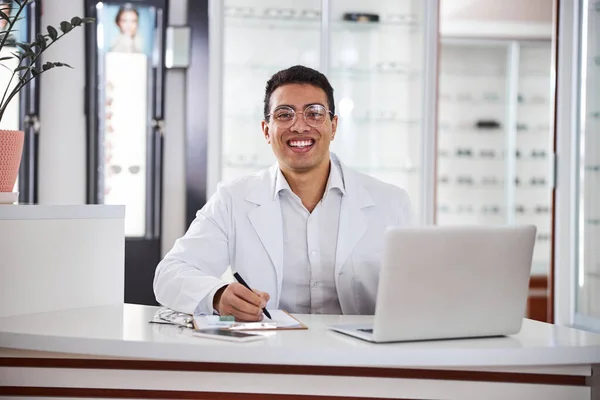 Sorridente spettegolato optometrista seduto a tavola nel suo ufficio — Foto Stock