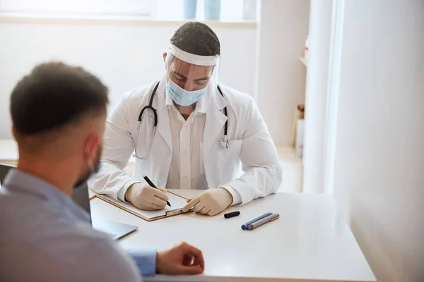 Guapo médico masculino escribiendo algo en papel blanco mientras escucha a su paciente en la habitación interior — Foto de Stock