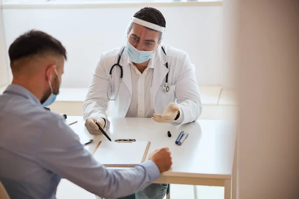 Médico masculino positivo en uniforme blanco con estetoscopio sentado en el escritorio mientras habla con un hombre con camisa azul — Foto de Stock