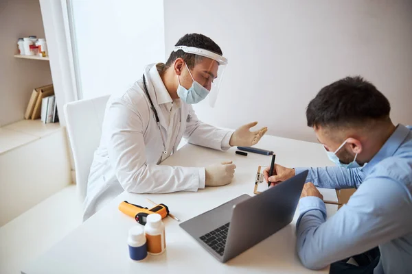 Terapeuta guapo profesional hablando con el hombre de camisa azul mientras escribe algo cerca del médico — Foto de Stock