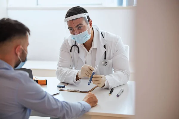 Hombre guapo en uniforme blanco y máscara protectora sentado a la mesa y mostrando herramienta para el hombre — Foto de Stock