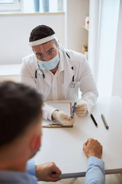 Zelfverzekerde arts in wit uniform en beschermend masker zittend aan het bureau met speciaal gereedschap in de hand — Stockfoto