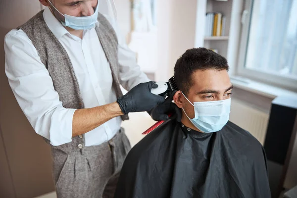 Красивий чоловік в чорному пігирі дивиться в сторону, сидячи в чоловічому салоні під час перукарні — стокове фото