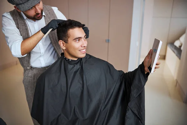 Pohledný muž v černém peignoir sedí v salonu krásy, zatímco holič tvorby styling s vlasy — Stock fotografie