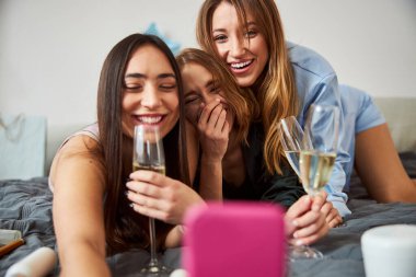 Şampanya kadehleriyle kendilerini yatakta fotoğraflayan neşeli kadınlar.