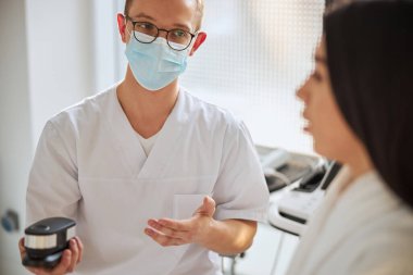 Gözlüklü doktor, siyah saçlı bir kadına tıbbi bir cihaz gösteriyor.