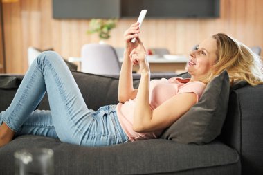 Neşeli kadın kanepede uzanıyor ve evde cep telefonu kullanıyor.