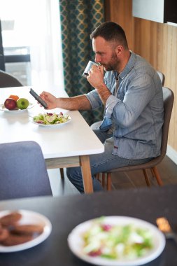 Sakallı adam akıllı telefon kullanıyor ve evde kahve içiyor.