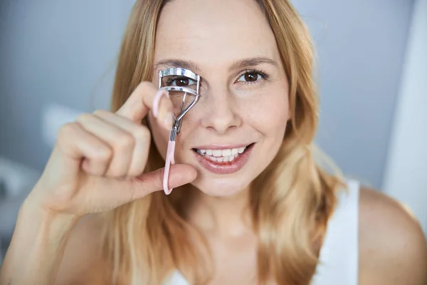 Cheerful young woman using eyelash curler at home