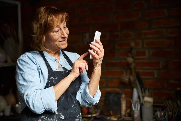 Χαρούμενη ηλικιωμένη γυναίκα που χρησιμοποιεί κινητό τηλέφωνο σε εργαστήριο κεραμικής — Φωτογραφία Αρχείου