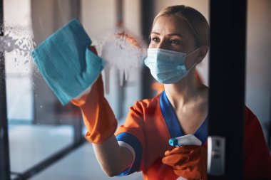 Maskeli ve lastik eldivenli bir kadın ofis yüzeyini temizliyor.