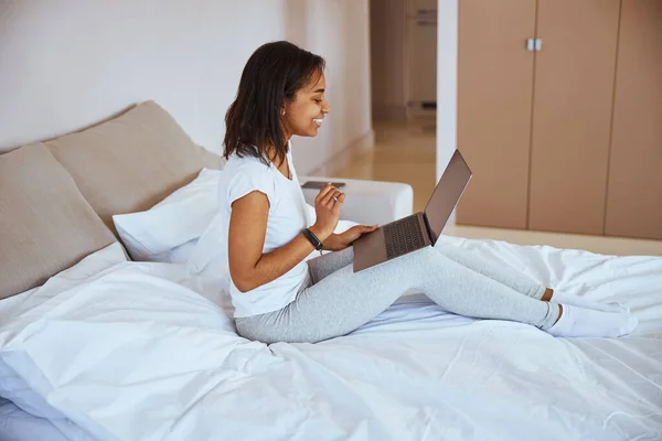 Χαρούμενη νεαρή γυναίκα που χρησιμοποιεί σύγχρονο φορητό υπολογιστή στο υπνοδωμάτιο — Φωτογραφία Αρχείου